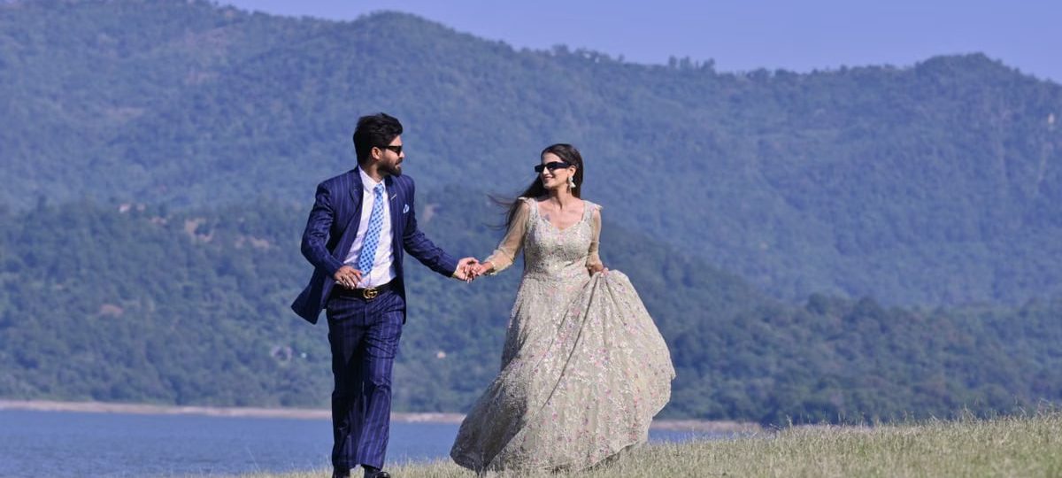 A couple in elegant wedding attire walking hand in hand by a picturesque riverbank in Una, India.